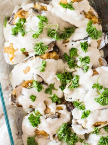 A glass dish filled with baked oysters topped with creamy sauce and garnished with fresh parsley. Lemon slices and oyster shells are placed nearby on a wooden surface, along with garlic cloves.