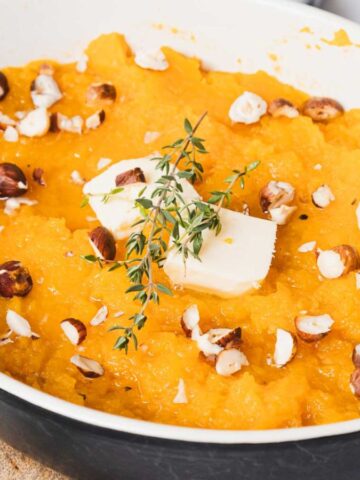 A dish filled with mashed sweet potatoes topped with chopped nuts, a pat of butter, and a sprig of thyme. The dish is placed on a wooden board, with a few thyme sprigs scattered around.