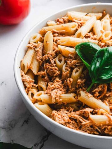 A bowl of penne pasta topped with shredded meat and garnished with fresh basil leaves. A fork is placed in the bowl. Red tomatoes and additional fresh basil leaves are on the marble surface nearby.