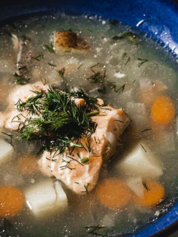A blue bowl contains a soup with chunks of fish, carrots, and potatoes, garnished with fresh dill. The soup appears to have a clear broth base.