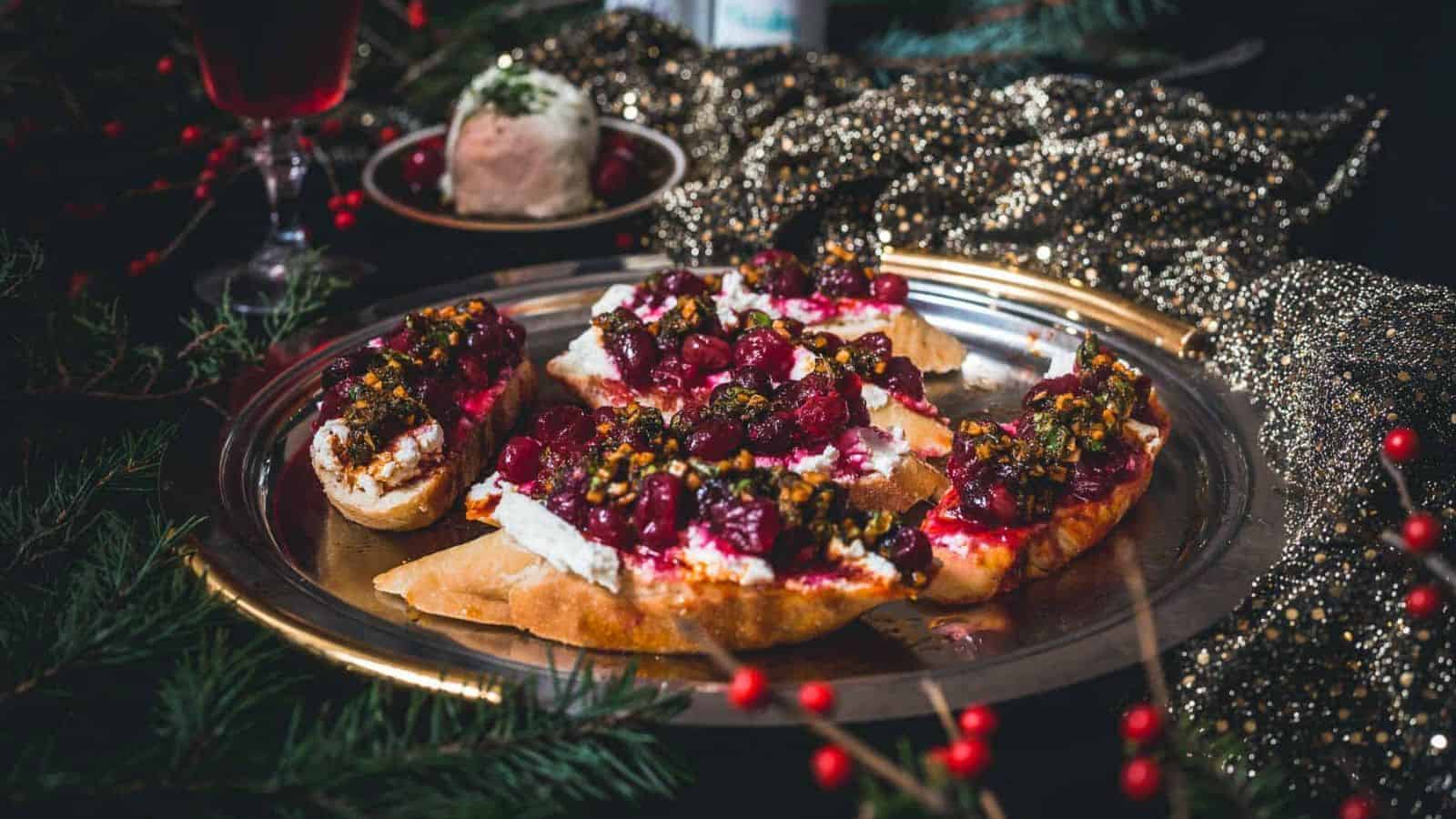 A festive platter features toasted bread topped with cream cheese, roasted cranberries, and a relish-like topping. The plate is decorated with dark, glittery fabric, surrounded by greenery and red berries, creating a holiday atmosphere.