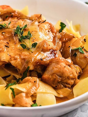 A white bowl holds a savory pappardelle pasta dish with braised chicken and mushrooms, reminiscent of French onion flavors, garnished with fresh herbs. A fork rests on a folded beige napkin beside the bowl, inviting you to indulge in this skillet-inspired creation.
