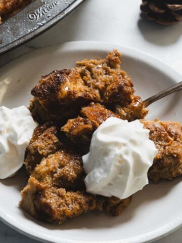 A plate of bread pudding topped with two dollops of whipped cream. A fork rests on the plate. In the background, a baking dish with more bread pudding is partially visible on a marble surface. Decorative items are placed around.