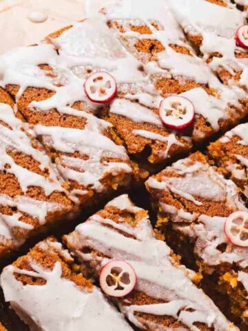 A rectangular loaf cake, sliced into pieces, is drizzled with white icing and topped with thinly sliced cranberries. The cake rests on a wooden surface.
