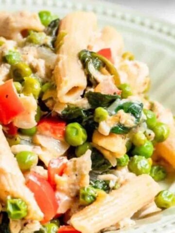 A plate of pasta salad featuring penne, peas, red bell peppers, spinach, and shredded chicken. The ingredients are mixed together on a light-colored, scalloped-edged plate, set against a plain background.
