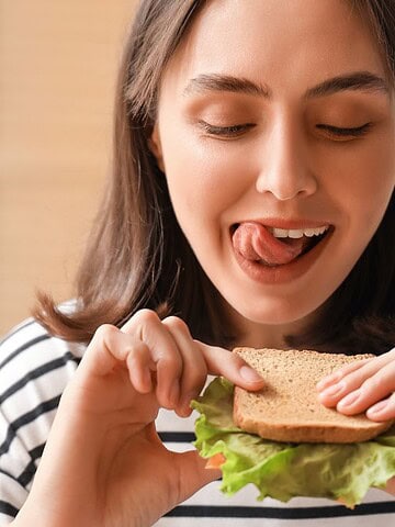 A woman in a striped shirt is holding one of her favorite sandwich combinations, complete with fresh lettuce and crispy toast. Smiling, she appears ready to take a bite, while the background remains softly blurred.