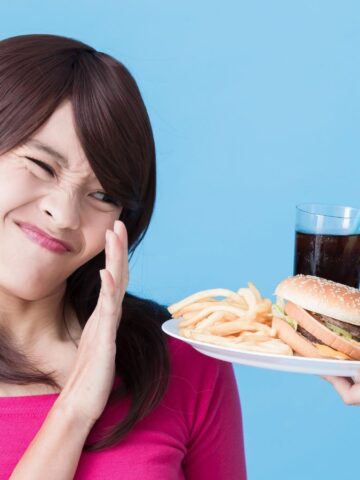 A woman in a pink shirt frowns and raises her hand to refuse a plate of classic American foods, including a burger, fries, a pink donut, and a soda. The background is blue.