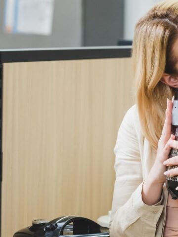 A person with blonde hair is in an office setting, smiling and embracing a large slow cooker snugly. The background features light wooden partition walls and some documents pinned on them, hinting at delicious soups and stews waiting to be made.
