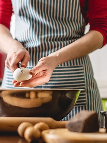 A person in a striped apron is breaking an egg into a large metal bowl in a kitchen, preparing quick holiday dishes. Various baking ingredients and utensils, including a rolling pin, milk, and chocolate, are on the wooden countertop.