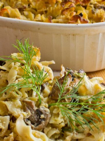 A plate of noodle casserole garnished with dill is placed on a wooden board next to a larger dish of the same casserole. The dish contains noodles, mushrooms, and a baked topping. The casserole is in a white ceramic container.