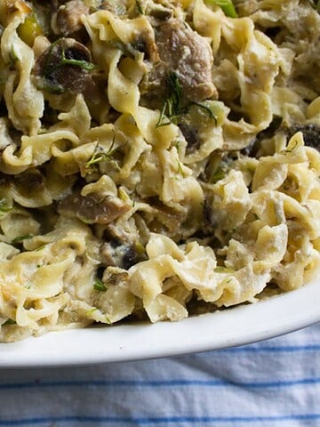 A white dish filled with creamy pasta topped with mushrooms and herbs is placed on a blue and white striped cloth.