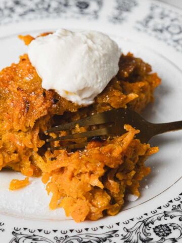 A slice of carrot cake on a decorative black and white plate, topped with a dollop of cream. A fork is inserted into the cake, which is set on a marble surface with part of a ceramic cup visible in the background.