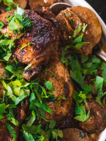 A roasted chicken garnished with fresh parsley is on a plate, surrounded by roasted potatoes. A bowl of white sauce is visible in the background. A silver serving spoon is partially tucked under the chicken.