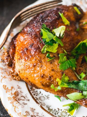 A cooked chicken leg with crispy skin is served on a decorative plate alongside roasted potatoes. The dish is garnished with fresh green herbs. A fork rests partially on the plate.