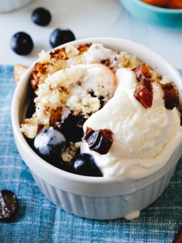 A dessert in a white ramekin contains crumbled cake with blueberries and a scoop of vanilla ice cream. A fork is placed on a blue cloth, surrounded by fresh oranges and prunes. A small bowl with orange slices is partially visible in the background.