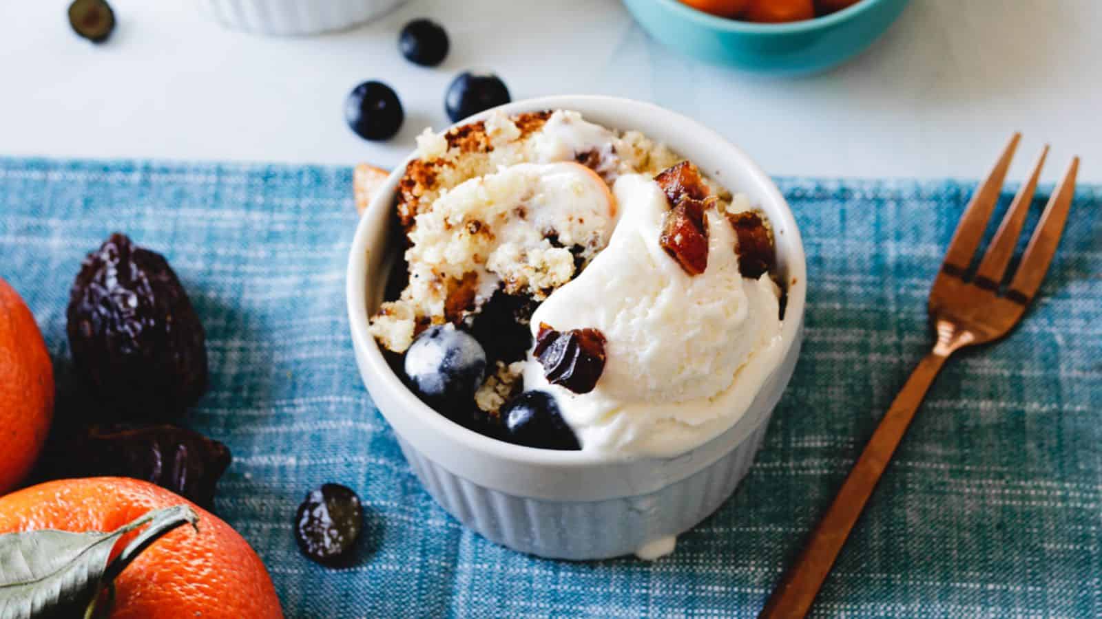 A bowl of dessert features a scoop of vanilla ice cream topped with pieces of chopped dates. Surrounding the ice cream are slices of yellow and purple fruits, along with dark grapes. A spoon rests in the bowl.