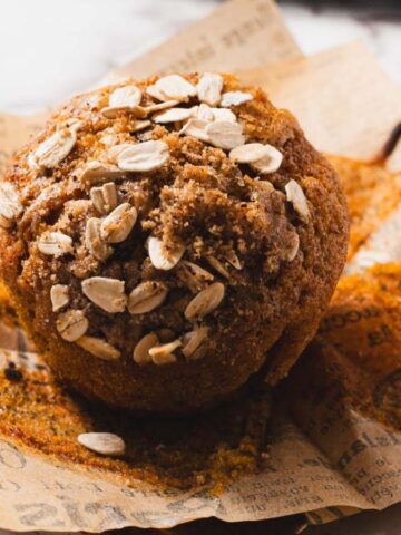 A close-up of a muffin topped with oats, resting on crumpled brown paper with printed text. The muffin is set on a marble surface.
