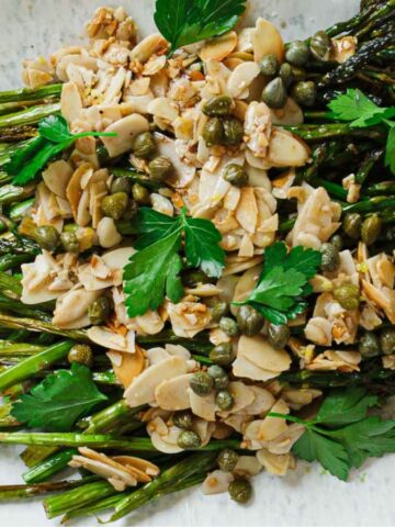 A plate of roasted asparagus topped with almond slices, capers, and parsley. Two lemon wedges are placed on either side of the asparagus. The dish is presented on an oval white plate.