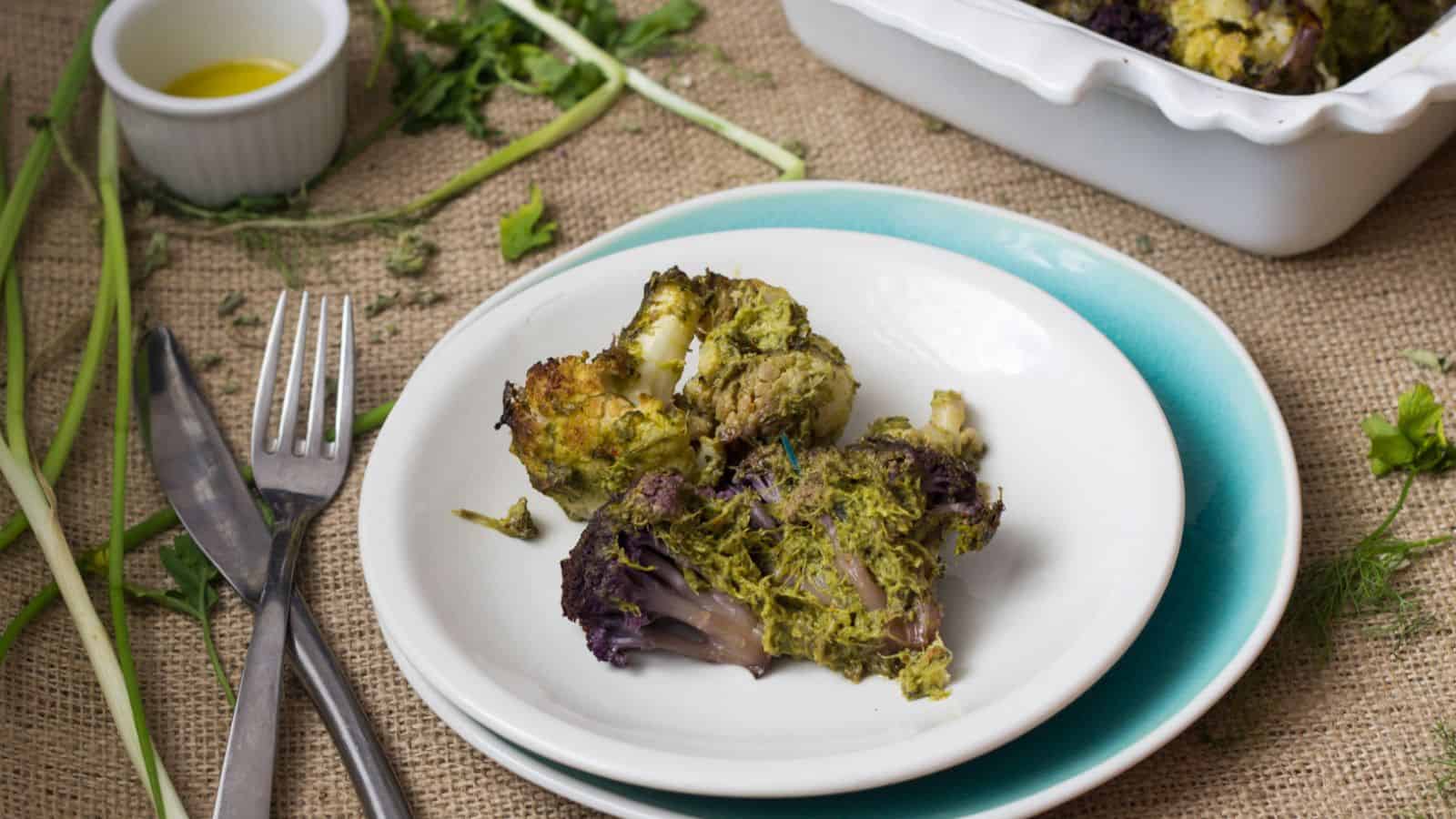 A baked dish with roasted cauliflower florets covered in a green herb sauce is displayed in a white ceramic baking dish. A sprig of fresh parsley garnishes the top. A small bowl of olive oil and a few scallions are placed beside the dish.