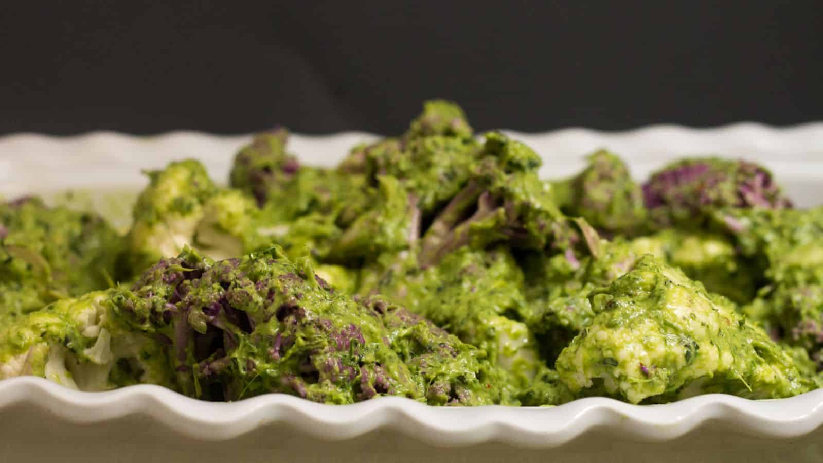 A baked dish with roasted cauliflower florets covered in a green herb sauce is displayed in a white ceramic baking dish. A sprig of fresh parsley garnishes the top. A small bowl of olive oil and a few scallions are placed beside the dish.