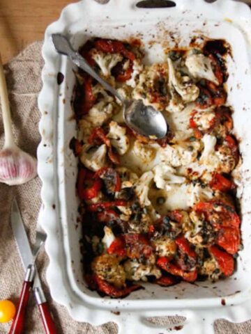 A comforting roasted vegetable dish in a white baking tray evokes memories of Grandma's Kitchen. It features cauliflower and tomatoes, sprinkled with herbs. A spoon rests in the tray, surrounded by a small bowl of capers, a garlic bulb, cherry tomatoes, and serving utensils on a cloth.