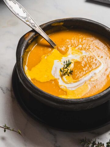 A bowl of creamy orange soup is placed on a black plate, garnished with a swirl of cream and a sprig of herb. A spoon rests inside the bowl. Small sprigs of thyme are scattered on the marble surface nearby.