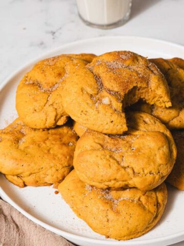 A plate of pumpkin cookies with sugar and cinnamon on top is placed on a marble surface. A bite is taken from one cookie. Two cinnamon sticks and a glass of milk are in the background, along with a beige napkin.