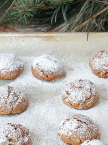 A baking sheet with numerous round cookies dusted with powdered sugar. Pine branches are visible in the background, adding a festive touch.
