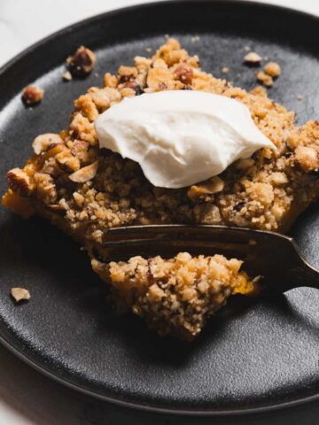 A slice of crumble dessert topped with a dollop of cream sits on a black plate. A fork rests on the plate, with crumbs scattered around. The dessert contains visible nuts and a crumbly texture.