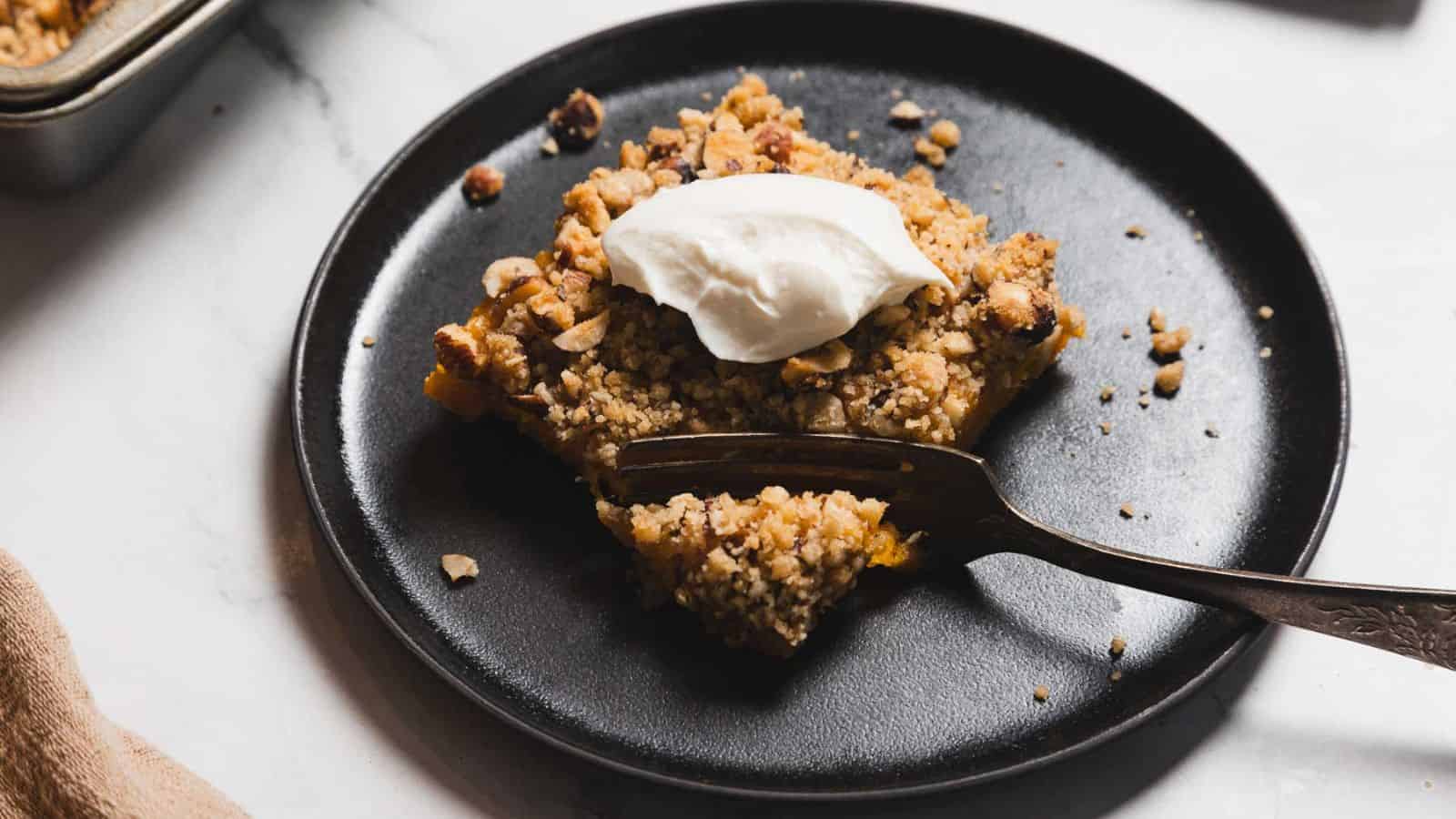 A slice of crumble dessert topped with a dollop of cream sits on a black plate. A fork rests on the plate, with crumbs scattered around. The dessert contains visible nuts and a crumbly texture.