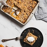 A baking pan with sliced pumpkin coffee cake topped with crumbly streusel evokes cozy autumn vibes. A piece is served on a black plate with a dollop of cream, reminiscent of sweet potato casserole with hazelnuts. A spoon rests in the pan, and a fork lies beside the plate on a marble surface.