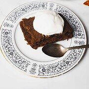 A slice of dark brown crockpot gingerbread cake topped with white cream sits on a decorative white plate with black floral patterns. A silver spoon rests beside the cake. Another plate with a similar cake slice is visible in the background. A pine cone is on the table.