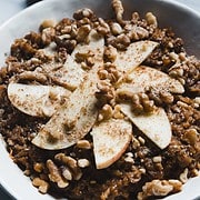 A bowl of slow cooker gingerbread oatmeal topped with sliced apples and walnuts, sprinkled with cinnamon. The dish is served on a white plate. A pine cone and a cinnamon stick are blurred in the background on a marble surface.