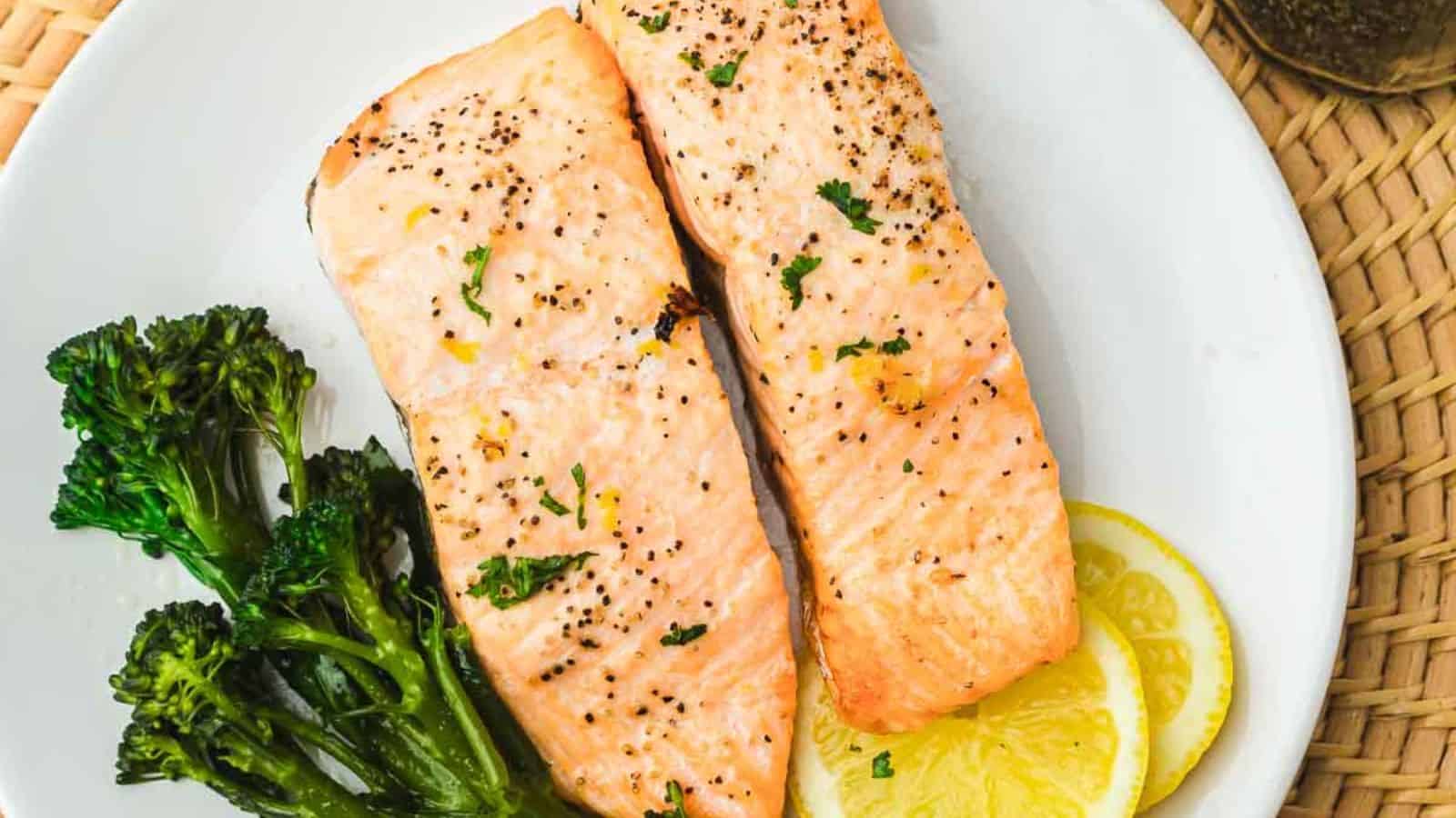 A white plate with two seasoned salmon fillets garnished with herbs. Broccolini is placed beside the salmon, and lemon slices are underneath. The plate is set on a wicker surface.