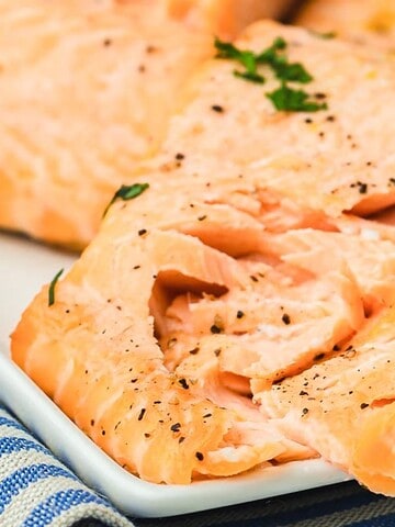 Close-up of a plate with cooked salmon fillets garnished with herbs and black pepper. A lemon wedge is placed next to the salmon. The plate is on a folded, striped cloth.