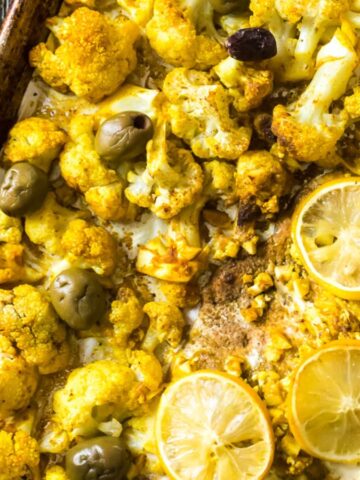A tray of roasted cauliflower with sliced lemons, olives, and spices. Nearby are small bowls containing green olives and a brown sauce, set on a burlap cloth.