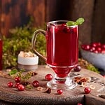 A glass mug filled with vibrant spiced cranberry juice garnished with mint and cranberries sits on a wooden board. Surrounding it are whole cranberries, star anise, and lush greenery, with a small bowl of cranberries in the background.