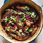 A large, oval baking dish contains a Festive Chicken with crispy, darkened skin. It is garnished with fresh parsley and pomegranate seeds, adding bright colors and textures. The dish rests on a light-colored, marble surface.