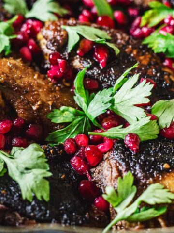 Close-up of a dish featuring browned meat with charred edges, topped with fresh green parsley and vibrant red pomegranate seeds. The ingredients are arranged in a rustic, deep dish.
