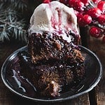 A slice of rich chocolate cake topped with melting vanilla ice cream and berry sauce sits on a dark plate. Red berries and pine branches are in the background, alongside a glass of spiced cranberry juice, adding a festive touch.