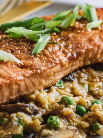 Grilled salmon fillet topped with chopped green onions, served on a bed of creamy risotto mixed with peas and mushrooms, on a gray plate with gold-colored cutlery.