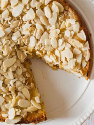A round cake topped with sliced almonds on a white plate, with a slice missing. Beside the plate, there is a small dish containing apple slices and a whole red apple on a white wooden surface.