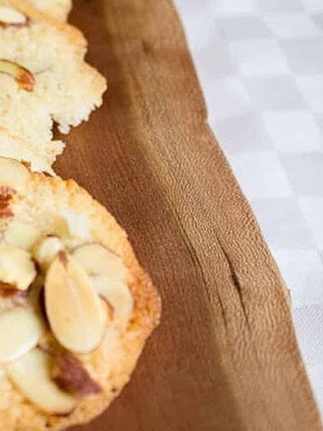 Four almond cookies with sliced almonds on top are placed on a wooden board. The cookies are arranged in a straight line.