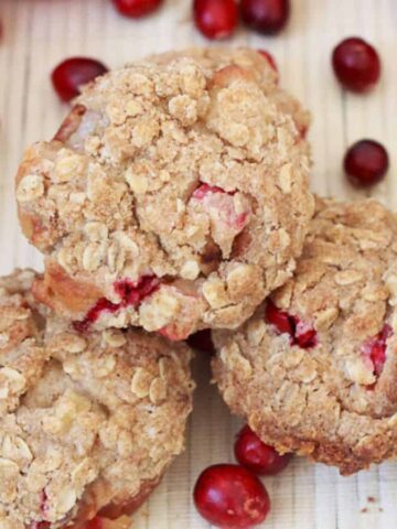 An arrangement of three oatmeal cranberry muffins surrounded by fresh apples and cranberries on a light-colored surface. The muffins have a crumbly texture, and the fruits are scattered around as decorative elements.