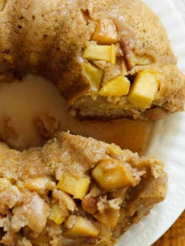 A close-up of a bundt cake on a white plate, featuring chunks of apple embedded in the cake. A slice has been removed, and a metal cake server with cut-out patterns rests beside it on a wooden table.