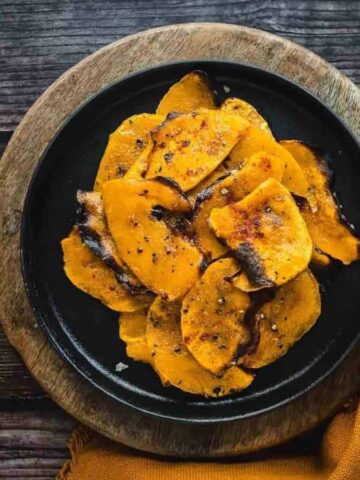 A black plate with a serving of roasted butternut squash slices seasoned with herbs. The plate is on a round wooden board with a yellow cloth beside it. Small pumpkins and a decorative bowl are on the left, all on a rustic wooden table.