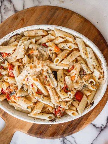 A white bowl filled with creamy penne pasta mixed with chunks of tomatoes and topped with herbs, placed on a round wooden board. The background features a marble surface scattered with whole tomatoes and blood orange halves.