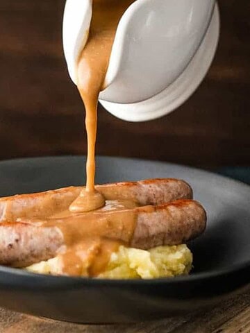A black plate with mashed potatoes topped with two sausages. Gravy is being poured from a white gravy boat over the sausages. The plate is on a wooden board, and a napkin is partially visible on the left side.
