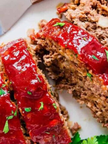 Close-up of slices of meatloaf topped with a red glaze, garnished with parsley, on a white plate. A metal spatula is visible next to the meatloaf.