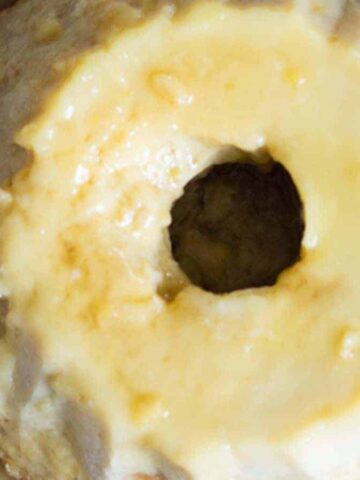 A bundt cake with a smooth, light yellow glaze sits on a white plate. The cake has a central hole, and the glaze appears drizzled over the top, creating a glossy surface.